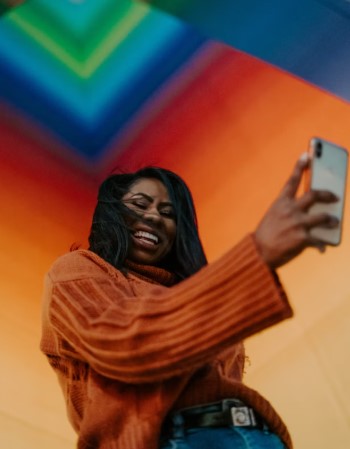 A young woman wearing an orange jumper and jeans standing in a colouful room and holding her smartphone in a good position to record a 'selfie' video.